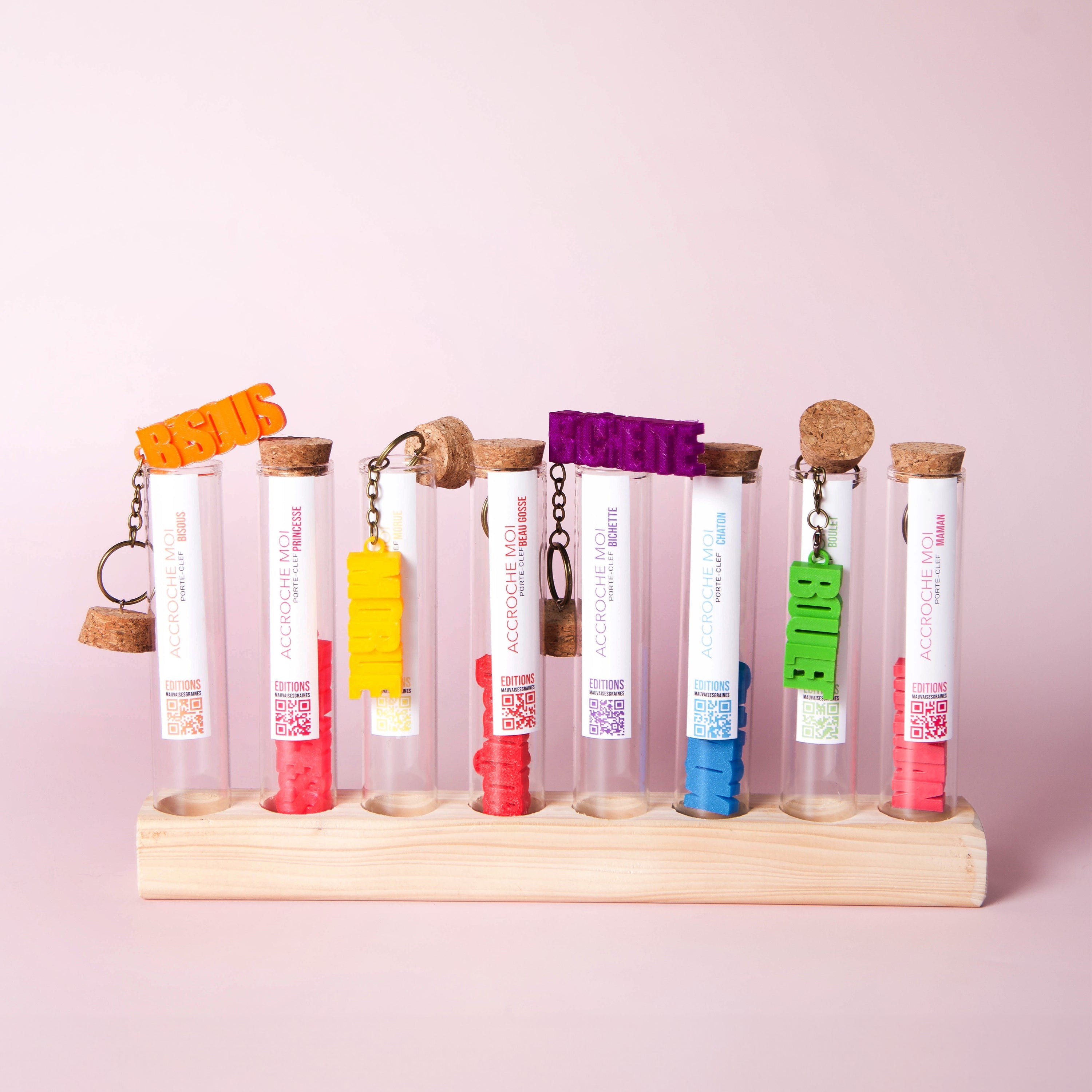 Série de porte-clés à message colorés, présentés dans des tubes à essai alignés sur un support en bois.